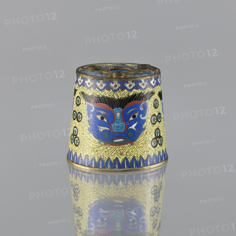 Sawdust container, cloisonné enamel, Chinese, Qing dynasty 1644–1911, 19th century. Vessel, height 8.5 cm, diameter 9.7 cm. Decoration on a yellow background: four masks against a grid background; motifs include beasts fantasy, tiger symbol, meander ornament, base with four inserts., A small conical metal object sits on a reflective surface against a plain gray background, showing a vibrant decorative face motif in bright blue with red and turquoise accents and black hairlike detailing, set on a yellow ground with an intricate geometric pattern; the piece has bands of deep blue along the top and bottom edges, small circular green and black medallions on the sides, areas of metallic gold outlining and trim, and a few worn or darkened spots on the metal rim, with the entire object mirrored in the surface below.