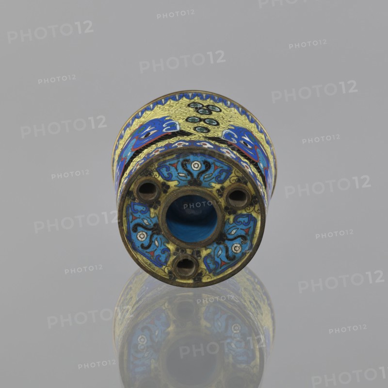 Sawdust container, gift, Chinese, Qing dynasty 1644–1911, cloisonné enamel vessel decorated with fantasy beasts, meander ornament, and tiger symbol; base with four inserts. Decoration: on a yellow background, four masks against a grid pattern. 19th century, China. Height 8.5 cm; diameter 9.7 cm., A cylindrical decorative object lies horizontally against a neutral gray background, showing its circular end facing the viewer and a reflective surface beneath that creates a mirrored image. The object has intricate enamel-like patterns in vivid blue, yellow, turquoise, black, white, red, and green, with stylized floral and abstract motifs wrapping around its body. The visible end has a central hollow opening surrounded by a ring and several smaller circular holes, all framed by ornate scrolling designs in blue and black on a yellow background with small white accents. The upper band shows repeating decorative elements in blue and yellow with touches of red and green, and the metal edges have...
