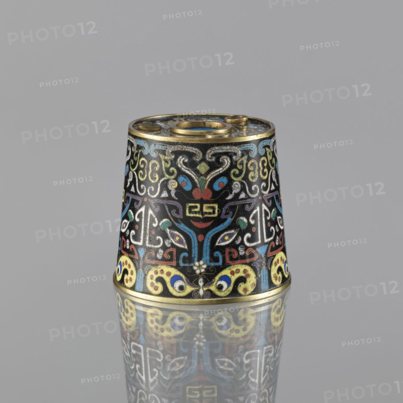 Sawdust container, gift, Paderewski, Ignacy Jan 1860-1941, Chinese art, Chinese, Qing dynasty 1644–1911, cloisonné enamel, fantasy beasts, cicadas, base with four inserts, 19th century, China, vessel; height 8.5 cm, diameter 9.7 cm, A small conical metal object sits upright on a reflective surface, showing a colorful enamel pattern against a black background with a gold rim at the top and bottom. The surface features ornate swirling motifs and stylized shapes in colors including blue, red, yellow, green, white, and purple. The design includes symmetrical, mask-like forms and curving decorative elements that repeat around the object. The reflection of the object is visible on the surface below, mirroring the same colors and patterns. The background is a plain gray.
