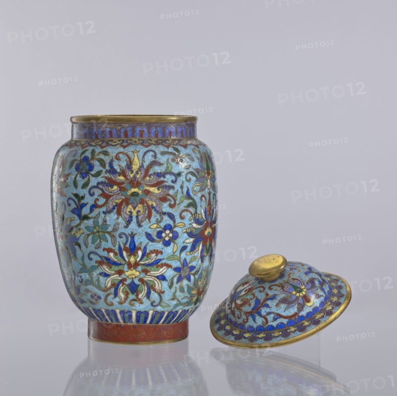 Vase with lid, Chinese, Qing dynasty 1644–1911, cloisonné enamel vessel with floral and plant ornaments including floral tendrils and lotus tendril motifs, decorative flowers; 18th–19th century, China; height 23 cm, diameter 14 cm., An ornate lidded jar and its lid sit against a pale gray background; the jar has a rounded body with a narrow rim and a separate domed lid positioned on its side nearby. Both pieces display an intricate floral pattern on a light blue ground, featuring flowers and scrolling foliage in colors including dark blue, red, green, and gold, with gold outlining and accents throughout. The rim and foot of the jar and the knob and edge of the lid show metallic gold tones, and there is a band of deep blue decoration near the base. The surface has a glossy finish that produces subtle reflections on the smooth tabletop, and the overall color palette includes light blue, dark blue, red, green, and gold.