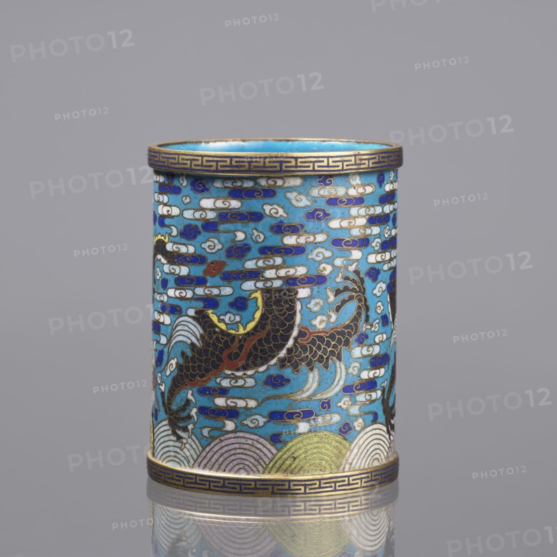Brush vessel; Chinese; Qing dynasty 1644–1911; cloisonné enamel decorated with dragons, clouds, waves, claws, and meander ornament; turquoise interior; 19th century; height 12.7 cm, diameter 9.8 cm., A cylindrical decorative object with a glossy surface featuring a predominantly turquoise blue background, decorated with a pattern of cloud-like shapes in white and deep blue, and several stylized fish or dragon-like creatures in dark brown and black with gold and red accents; the top and bottom rims have a narrow band with a repeating geometric motif in gold and dark blue, and the lower edge shows layered wave patterns in pale green, cream, and muted gold; the overall color palette includes turquoise, deep blue, white, dark brown, black, gold, red, pale green, and cream, and the object is shown against a smooth gray background with a faint reflective surface beneath it.