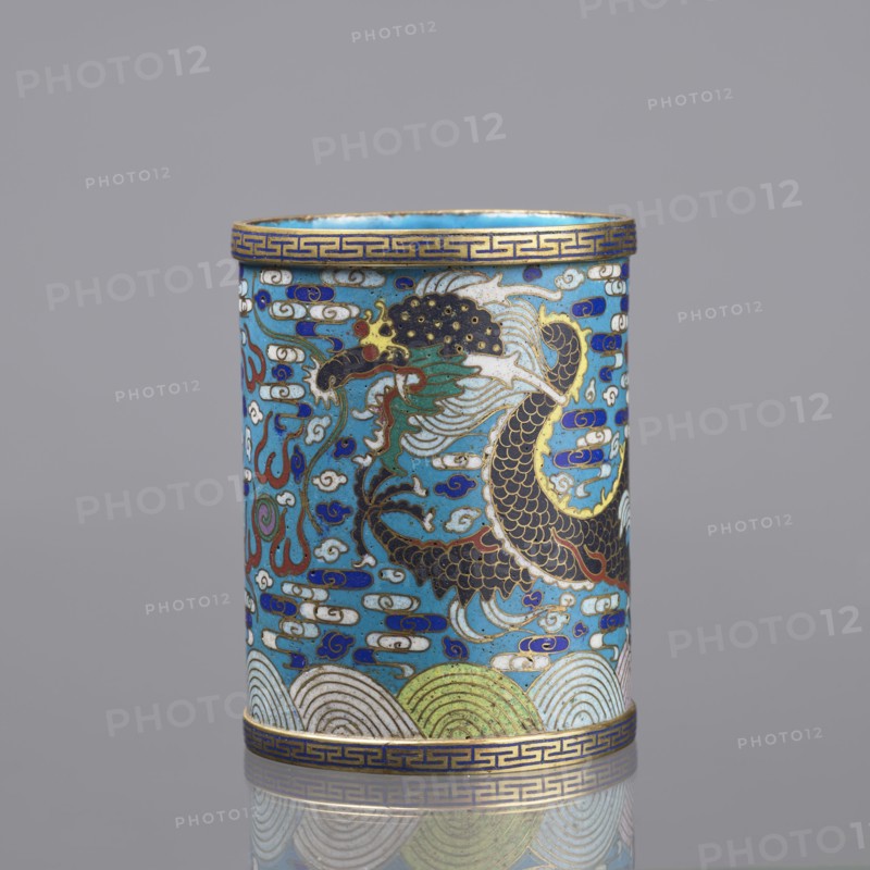 Brush vessel, 19th century, China; Qing dynasty; cloisonné enamel; subject: dragons, clouds, waves, claws, meander ornament; function: brush vessel; turquoise interior; height 12.7 cm, diameter 9.8 cm., A cylindrical decorative object shown upright with a glossy surface, featuring a dominant turquoise blue background and detailed cloisonné-style enamel work. The central area displays a stylized dragon or serpentine creature rendered in black, dark brown, green, white, red, and gold outlines, with flowing mane-like shapes and scale patterns. Surrounding the creature are swirling cloud and wave motifs in white, blue, dark blue, and touches of red and green. The top and bottom rims have a repeating geometric border in gold on a blue band. The lower section includes scalloped wave patterns in pale green, white, and light blue with gold accents. The overall palette includes turquoise blue, dark blue, white, black, gold, green, red, and brown, and the piece sits against a neutral gray backgr...