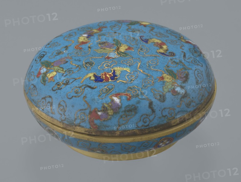 Round box with lid, cloisonné enamel, Chinese, Qing dynasty 1644–1911, 19th century. Cylindrical flattened shape on a separate ring foot with a slightly convex lid. Entire surface decorated with multi-colored lotus tendrils on a turquoise background. Lid shows three freely placed fruits — a peach, a pomegranate, and a lemon — accompanied by bats; motif interpreted as wishing many happiness, long life, and many sons. Asymmetrical composition set against cloud forms made of bent wire; same motif appears in the box's interior storage section, which is also decorated with colorful medallions single, paired, or grouped in threes featuring various Chinese plant ornaments. Rounded edges of lid and body, lower edge of foot and bottom are gilded. Bottom and entire interior surface covered with turquoise enamel. Container. Height 4.6 cm; diameter 9.8 cm., A rounded lidded container with a domed top and a circular base, predominantly bright blue in color with a gold rim and gold decorative outlin...