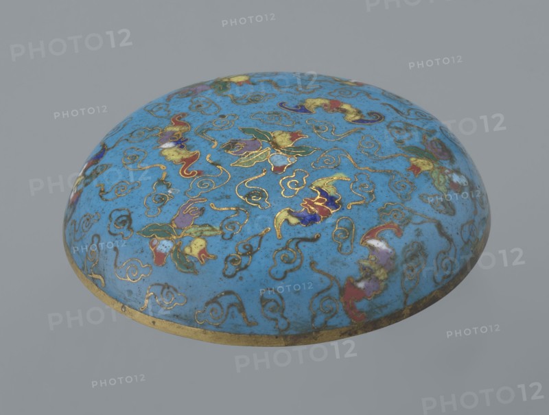 Round box with a lid, Chinese, Qing 1644–1911, cloisonné enamel, cylindrical flattened shape on a separate ring foot with a slightly convex lid, decorated overall with multi-colored lotus tendrils on a turquoise background. Lid shows three fruits peach, pomegranate, lemon with accompanying bats—an auspicious motif meaning many happiness, long life, and many sons. Asymmetrical composition set against a cloud background formed by bent wire; same motif appears in the box’s storage section, which is also decorated with colorful medallions single, pairs, or threes featuring various Chinese plant ornaments. Rounded edge of lid and storage section, lower edge of foot and its bottom are gilded. Bottom and entire interior covered with turquoise enamel. 19th century, China; container; height 4.6 cm, diameter 9.8 cm., A rounded dome-shaped object with a glossy blue surface decorated with a pattern of swirling line motifs and floral-like shapes outlined in metallic gold, with additional colors inc...