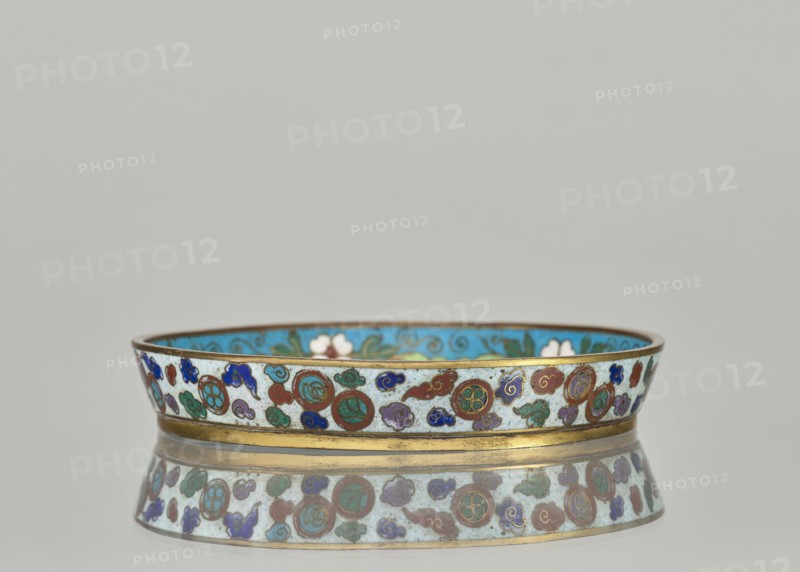 Plate, cloisonné enamel dish with brim, 19th century China Qing dynasty. Materials: cloisonné enamel. Decoration: plant ornaments and tiger motif symbol. Dimensions: height 2.4 cm, diameter 14.2 cm., A low, round decorative bowl with a thin gold rim and gold band near its base sits on a reflective surface, showing a mirrored image below. The exterior of the bowl features a repeating pattern of circular medallions and cloud-like motifs in colors including turquoise, blue, green, red, purple, and white against a pale background. The interior visible at the rim appears bright turquoise with floral shapes in white and green. The overall scene includes neutral gray background tones.