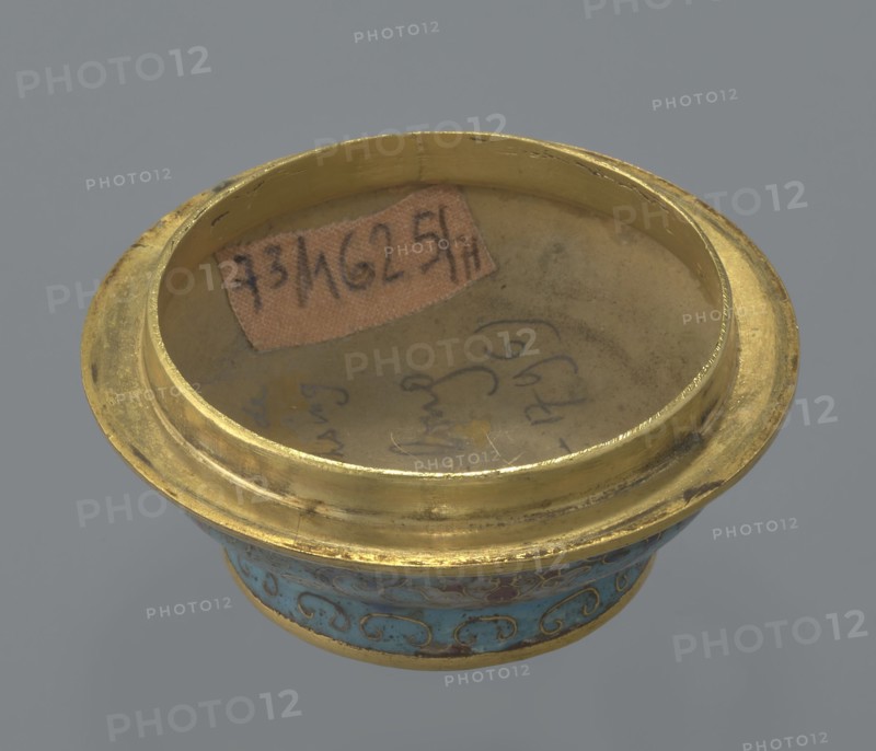 Box with a lid, Chinese, Qing dynasty, Qianlong period 1736–1795, cloisonné enamel, cylindrical spheroidal shape on a separate ring foot, slightly convex lid. Entire surface decorated with multi-coloured lotus tendril on a turquoise ground; foot bears a band with curling silkworm-larva motif. Rounded edge of lid, rim of box, lower edge of foot, interior and bottom gilded. Bottom engraved with inscription made in the Qianlong era and workshop/guild mark yue. Pair with a similar item; two pieces differ slightly in shape, height, and ornamentation. 18th century, China. Container; height 4 cm, diameter 6.7 cm., A shallow circular metal vessel with a wide rim, showing a worn golden surface and a few small dark spots, sits against a gray background; the interior base has a muted beige tone with some faint smudging and a small irregular patch of brownish paper, and the exterior band near the base displays a decorative turquoise area with swirling gold patterns and touches of darker blue and g...