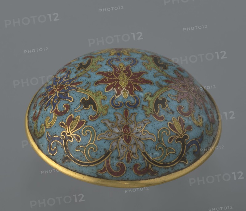 Cylindrical cloisonné enamel box with lid, spheroidal shape on a separate ring foot, slightly convex lid, decorated overall with multi-coloured lotus tendril ornament on a turquoise background. Foot bears a band with a curling silkworm-larva decorative motif. Rounded edge of lid, storage section, lower edge of foot, interior and bottom are gilded. Bottom bears an engraved signature made in the Qianlong era and a workshop/guild mark yue. Pair with a similar item that differs slightly in shape, height, and ornamentation. Qing dynasty, Qianlong period 18th century, China. Height 4 cm; diameter 6.7 cm. Materials: cloisonné enamel., A rounded ornamental object with a domed surface decorated in a light blue background and ornate floral and scroll patterns in gold outlines filled with colors including red, dark blue, green, purple, black, and muted beige, with a gold rim around the edge.