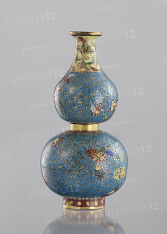 Double gourd-shaped vase, Chinese, Qing dynasty 1644–1911, cloisonné enamel decorative vessel. Decoration: flowers, butterflies, insects on a turquoise ground with a grid of circles; yellow neck. Twisted from two parts. 19th century. Height 14 cm, diameter 7 cm., A double-gourd shaped decorative object with a glossy surface showing a dominant blue color patterned with an intricate gold interlocking ring motif, accented with floral and butterfly-like designs in red, purple, yellow, and green, a narrow neck area that is lighter with pale yellow and green tones and similar floral accents, thin gold bands separating the two bulbous sections and forming the rim and base edge, a small section near the base showing a band of dark red with pale yellow circular spots, and a neutral light gray background.