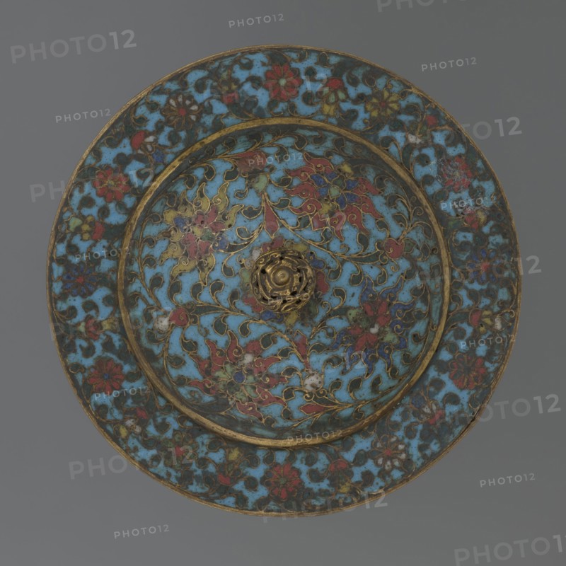 Tin with a lid, Chinese, Qing dynasty, Qianlong period, 18th century. Cylindrical tin with a wide plate-like collar extending beyond the vessel walls and slightly rising at the rim. Domed lid with a handle in the shape of a decorative knob with openwork relief: a dragon in clouds, its head directed toward a pearl at the top of the handle. Exterior of the vessel box and collar decorated in cloisonné enamel with a multicolored red, blue, green, yellow, white small-flowered ornament — branches with rosettes, buds, and leaves — on a turquoise background. Lid decorated with a lotus tendril of four geometrically arranged flowers and surrounding branches; a multi-petal rosette at the center under the metal handle, all on a turquoise ground. Bottom filled with a rich multicolored composition of elements reminiscent of Buddhist symbols — a vajra and Dharma wheel forming the inner central rosette, with red and blue ribbons between the symbols. Interior of box and lid ungilded of enamel; all inte...
