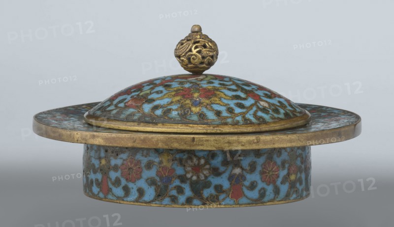 Tin with a lid, Chinese, Qing dynasty, Qianlong period, cloisonné enamel. Cylindrical tin with a wide plate-like collar extending beyond the walls and slightly rising at the rim. Domed lid with a handle in the form of an openwork relief dragon in clouds, head directed toward a pearl at the top of the handle. Exterior of vessel box and collar decorated with multicolored red, blue, green, yellow, white small-flower ornament — branches with rosettes, buds, and leaves — on a turquoise background. Lid decorated with a lotus tendril of four geometrically arranged flowers and surrounding branches; multi-petal rosette beneath the metal handle, all on a turquoise ground. Bottom filled with a rich multicolored composition of elements reminiscent of Buddhist symbols — a vajra and Dharma wheel forming the inner central rosette, with two-colored red and blue ribbons between the Buddhist symbols. Interior of box and lid unenameled and gilded. Possibly an element of religious ritual incense box. 18th...