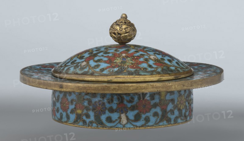 Tin with a lid, Paderewski, Ignacy Jan 1860–1941, Chinese, Qing 1644–1911, Qianlong period, cloisonné enamel. Cylindrical tin with a wide plate-like collar extending beyond the vessel walls, slightly rising at the rim. Domed lid with a handle shaped as a decorative openwork relief: a dragon in clouds, head directed toward a pearl atop the handle. Exterior box and collar decorated with a multicolored red, blue, green, yellow, white small-flowered ornament — branches with rosettes, buds, and leaves — on a turquoise background. Lid decorated with a lotus tendril of four geometrically arranged flowers and surrounding branches; center beneath the metal handle features a multi-petal rosette, also on turquoise. Bottom filled with a rich multicolored composition of elements reminiscent of Buddhist symbols — a vajra and Dharma wheel forming the inner central rosette, with two-colored red and blue ribbons between the symbols. Interior of box and lid lacks enamel decoration; all interior elements...