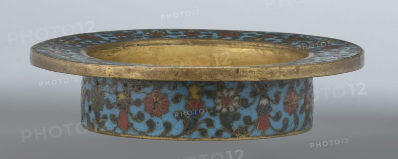Tin with a lid;, Chinese, Qing dynasty, Qianlong period, 18th century. Cloisonné enamel on tin; cylindrical container with a wide plate-like collar extending beyond the vessel walls and slightly rising at the rim; domed lid with a decorative openwork knob depicting a dragon in clouds facing a pearl. Exterior box and collar decorated with multicolored red, blue, green, yellow, white small-flowered ornament — branches with rosettes, buds, and leaves — on a turquoise background. Lid decorated with a lotus tendril composed of four geometrically arranged flowers and surrounding branches; a multi-petal rosette centered beneath the metal handle, also on turquoise. Bottom filled with a rich multicolored composition of elements reminiscent of Buddhist symbols a vajra and Dharma wheel forming the central rosette with two-colored red and blue ribbons between the symbols. Interior of box and lid gilt without enamel. Possibly used as a ritual incense box. Height 7.7 cm; diameter 15.1 cm., A shallow...