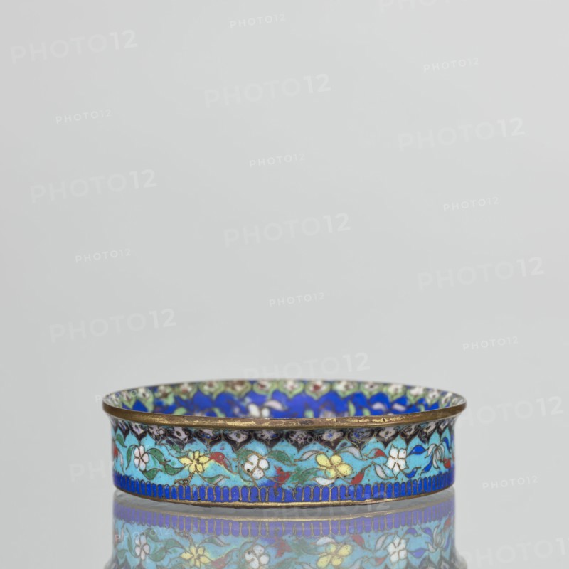 Bowl, 19th century, China. Cloisonné enamel decorative vessel, height 2.3 cm, diameter 9.4 cm. The round, flat-bottomed bowl with a low side wall widening toward the rim is decorated in the mirror with an ornamental composition on a light purple background arranged around a central black rosette. The composition includes four butterflies in white, red, and black, multicolored flowers including Buddha's fingers/Chinese lemon and surrounding leaves arranged in a circle. The inner wall is decorated with a floral tendril against a blue background composed of multicolored flowers similar to those in the mirror. A lambrequin ornament at the edge resembles the shape of rui heads. The rim is curved outward and gilded. The turquoise outer wall is filled with decoration similar to that on the inner wall. The underside of the bottom is covered with a single-color purple enamel. Pair with another bowl. Qing dynasty. Chinese., A shallow round metal dish with a decorative rim sits centered against a...