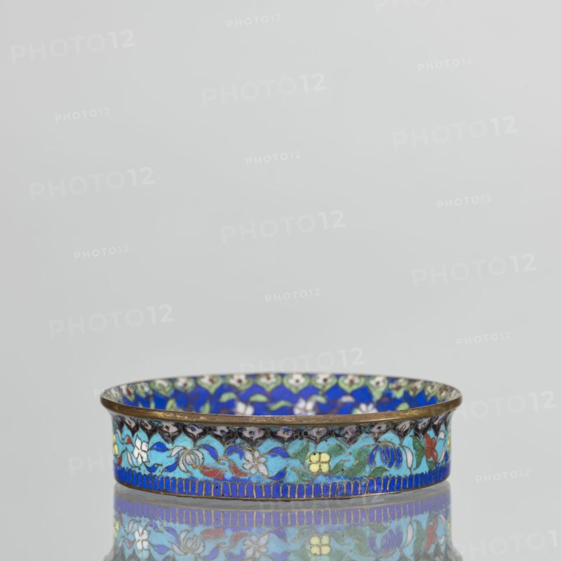 Bowl, gift, Chinese, Qing dynasty 1644–1911, cloisonné enamel decorative vessel, 19th century, China; vessel, height 2.3 cm, diameter 9.4 cm. The round, flat-bottomed bowl with a low side wall widening toward the rim is decorated in the mirror with an ornamental composition on a light purple background arranged around a central black rosette. The composition features four butterflies in white, red, and black, multicolored flowers including Buddha's fingers/Chinese lemon and surrounding leaves arranged in a circle. The inner wall is decorated with a floral tendril against a blue background with multicolored flowers similar to those in the mirror. A lambrequin ornament is visible at the edge, reminiscent of the shape of rui heads. The rim is curved outward and gilded. The turquoise outer wall is filled with decoration similar to that on the inner wall. The underside of the bottom is covered with a single-color purple enamel. This piece is a pair with another bowl., A shallow round dish w...