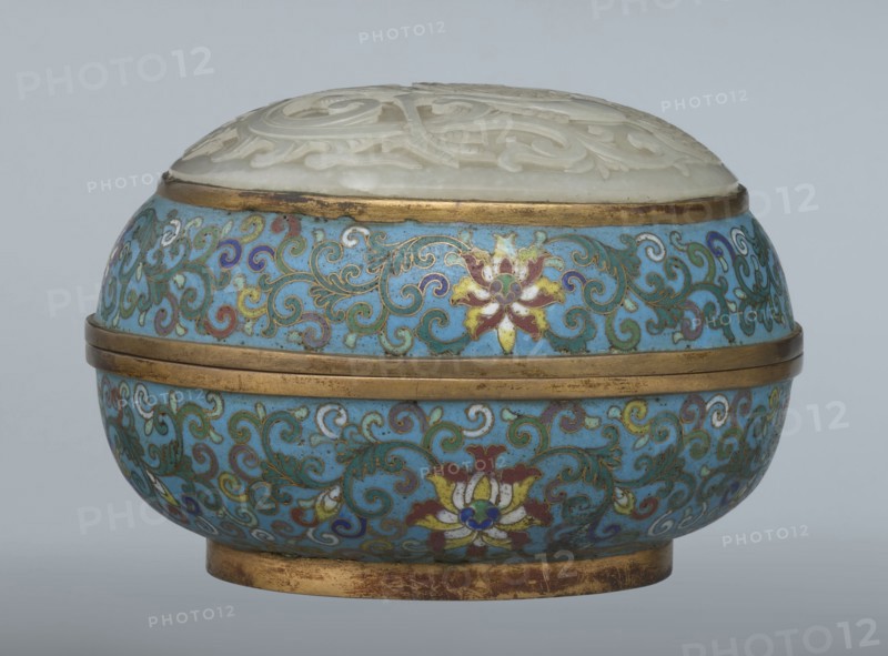 Box with a lid, Chinese, Qing dynasty 1644–1911, Qianlong period, cloisonné enamel; oval spheroidal box based on a ring foot, divided halfway into container and lid. Slightly convex bas-relief grayish jade plaque on the lid depicting a fenghuang phoenix or peacock with a sun among clouds; sun in upper left surrounded by a silk ribbon. Motifs and styling reference emblems from mandarin squares: peacock denoting third civil rank and sun referring to imperial authority. The jade plaque is a fine example of hard-stone workmanship. Rounded sides of container and lid decorated with multi-colored lotus tendril stripes on a turquoise ground. Interior filled with a uniform layer of turquoise enamel. Metal rings reinforcing the edges of the container, lid, and plaque attachment are gilded. 18th century, China. Height 8.2 cm; width 11.3 cm; diameter 12.2 cm., A rounded decorative container with a domed carved lid sits against a plain pale gray background; the lid appears off-white or cream with c...