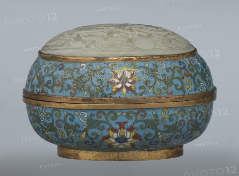 Box with a lid, cloisonné enamel, 18th century, China. Oval, spheroidal box on a ring foot, divided halfway into container and lid. Slightly convex bas-relief grayish jade plaque added to top of cover depicting a fenghuang phoenix or peacock with a sun among clouds; the sun is surrounded by a silk ribbon. Motifs and styling reference emblems on mandarin squares—peacock denoting third civilian rank and the sun referring to imperial authority. Rounded sides of container and lid decorated with a multicolored lotus tendril stripe on a turquoise ground. Interior filled with a uniform layer of turquoise enamel. Metal rings reinforcing edges of container, lid, and plaque attachment are gilded. Height 8.2 cm; width 11.3 cm; diameter 12.2 cm., A round decorative box with a domed lid carved from a pale, creamy material and an ornate body covered in a bright turquoise-blue enamel patterned with swirling vine motifs and stylized floral designs in colors including gold, green, red, white, and purpl...