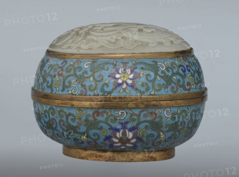 Box with a lid, Chinese, Qing dynasty, Qianlong period, cloisonné enamel on metal with a slightly convex bas-relief grayish jade plaque on the lid. Oval spheroidal box on a ring foot, divided halfway into container and lid. Jade plaque depicts a fenghuang phoenix or peacock with a sun among clouds; the sun is upper left surrounded by a silk ribbon. Motifs and styling reference emblems from mandarin squares: peacock indicating third civil rank and the sun referring to imperial authority. Plaque is a fine example of jade workmanship. Rounded sides of container and lid decorated with a multicolored lotus tendril band on a turquoise ground. Interior filled with uniform turquoise enamel. Metal rings reinforcing edges and plaque attachment are gilded. 18th century, China. Height 8.2 cm; width 11.3 cm; diameter 12.2 cm., A rounded decorative container with a domed carved lid sits against a plain pale gray background; the lid appears white and carved with raised swirling floral motifs and rest...