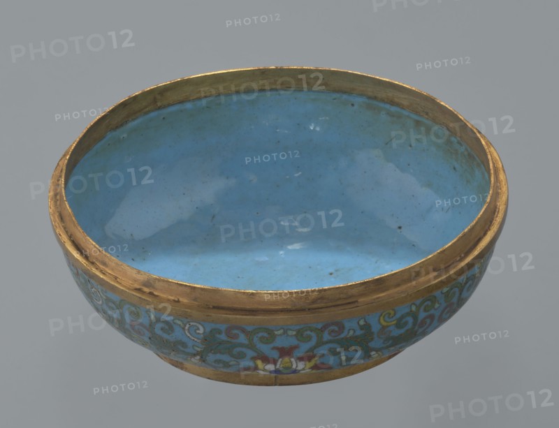Box with a lid. Oval, spheroidal box on a ring foot, divided halfway into container and lid. Cloisonné enamel with gilded metal reinforcing rings; interior filled with uniform turquoise enamel. Slightly convex bas-relief grayish jade plaque added to top of cover, depicting a fenghuang phoenix or peacock with a sun among clouds; sun is upper left, surrounded by a silk ribbon. Motifs and styling reference emblems from mandarin squares: peacock denoting third civilian rank, sun referring to imperial authority. Rounded sides of both container and lid decorated with multi-colored lotus tendril stripes on a turquoise ground. Exceptionally fine jade workmanship noted; early precursor cloisonné technique used semi-precious stones set in metal cells. 18th century, China. Height 8.2 cm; width 11.3 cm; diameter 12.2 cm. Creator, : Paderewski, Ignacy Jan 1860–1941., A shallow round bowl with a smooth glossy interior in shades of blue and turquoise, bordered by a worn metallic rim in gold tones; th...