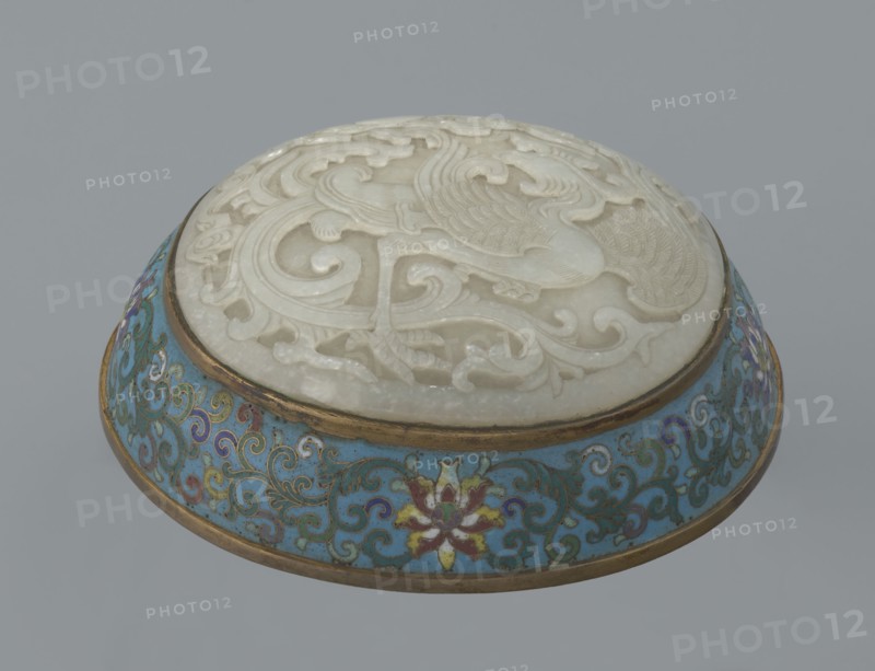 Box with a lid., Chinese, Qing dynasty, Qianlong period, 18th century. Cloisonné enamel on metal with a slightly convex bas-relief grayish jade plaque on the lid. Oval, spheroidal box based on a ring foot, divided halfway into container and lid. Lid plaque depicts a fenghuang phoenix or peacock with a sun among clouds; the sun is in the upper left surrounded by a silk ribbon. Motifs and styling reference mandarin square emblems peacock denoting third civilian rank; sun referring to imperial authority. Rounded sides of both container and lid decorated with multi-colored lotus tendril stripes on a turquoise ground. Interior filled with a uniform layer of turquoise enamel. Metal rings reinforcing the edges and the plaque attachment are gilded. Height 8.2 cm; width 11.3 cm; diameter 12.2 cm., A circular decorative object with a carved white top showing swirling raised patterns and bird-like shapes set into a shallow rounded base with a band of gold trim; the base features a turquoise backg...