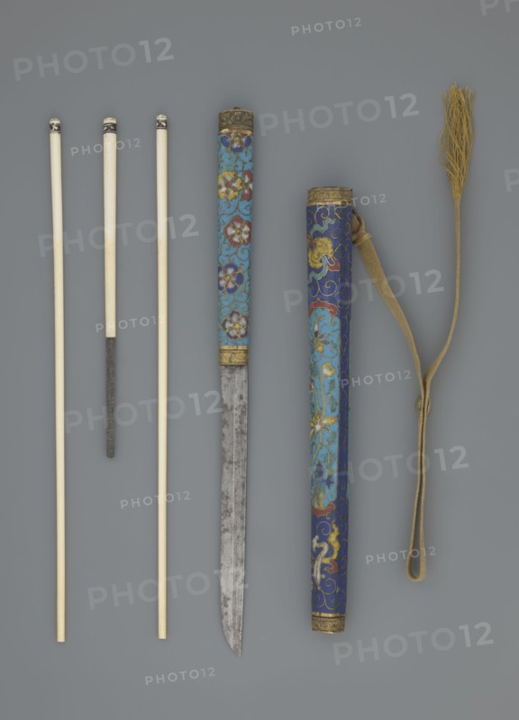 Travel cutlery set in case; Chinese, Qing dynasty 1644–1911, 19th century. Consists of: a Knife with cloisonné enamel handle and steel blade. b–c Ivory chopsticks, each decorated at the top with an inlaid black lacquer border ornament. d File decorated similarly to the chopsticks. e Case made of cloisonné enamel with bronze fittings engraved at top and bottom; interior lined with wood. Chopsticks and file held by a band decorated with a matching border; a narrow cotton-silk strap with fringes is attached. Case decorated with flowers in two reserves and plant tendrils in the background. Materials: cloisonné enamel, ivory, steel, bronze, lacquer. Function: travel cutlery. Length: 28 cm case. Iconography: plant motifs, swastika., A group of long slender objects arranged vertically on a gray background: a few pale ivory rods with smooth surfaces and darkened tips, one of which has a textured darker section partway down; a decorated cylindrical sheath and a matching scabbard both showing in...