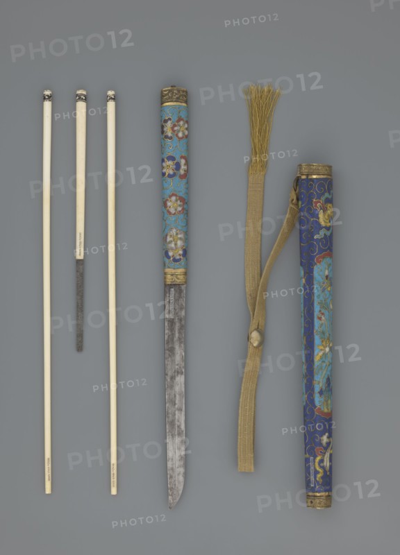 Travel cutlery set in case; Chinese, Qing dynasty 1644–1911, 19th century. Includes: a Knife with cloisonné enamel handle and steel blade. b–c Ivory chopsticks decorated with an inlaid black lacquer ornament at the top in the form of a border. d File decorated similarly to the chopsticks. e Case reaching 3/4 of the height of the cutlery; made of cloisonné enamel with bronze fittings engraved at top and bottom; interior lined with wood. Chopsticks and file are secured with a band decorated with a matching border; a narrow cotton-silk strap with fringes is attached. Case decorated with flowers in two reserves and plant tendrils in the background. Motifs include plants and a swastika. Cutlery; length 28 cm case., A group of thin, pale rods with dark tips arranged vertically on the left, a long narrow blade with a patinated metallic surface and a decorated cylindrical handle featuring floral patterns in blue, red, white, and gold near the center, a long woven strap in tan with a fringed en...