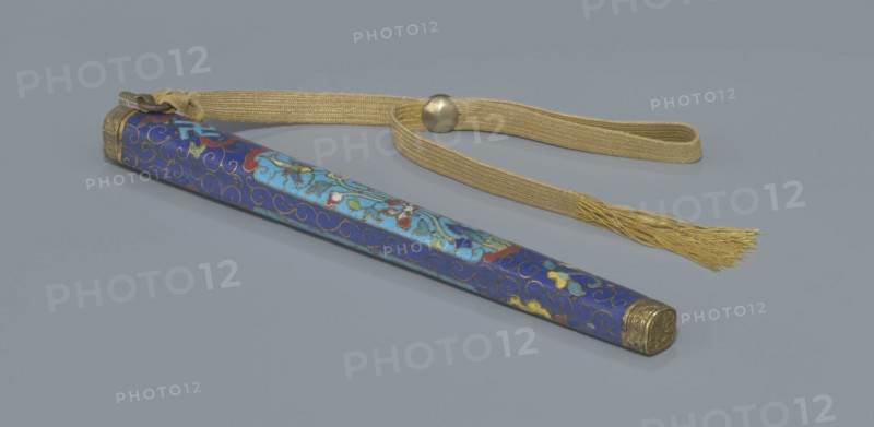 Travel cutlery set in case;, 19th century, China, Qing dynasty. Travel cutlery comprising: a Knife with cloisonné enamel handle and steel blade; b–c Ivory chopsticks decorated with an inlaid black lacquer border ornament at the top; d File decorated similarly to the chopsticks; e Case 28 cm length of cloisonné enamel with bronze fittings engraved at top and bottom, interior lined with wood. The chopsticks and file are held by a band decorated with a matching border; a narrow cotton-silk strap with fringes is attached. Case decorated with floral reserves and plant tendrils in the background. Motifs include plant designs and a swastika. Function: cutlery., A long slender sheath with a decorative blue surface featuring swirling gold outlines and floral-like motifs in lighter blue, red, green, yellow, and white rests on a flat grey background, with ornate metallic gold caps at each end; attached to one end is a woven gold-colored strap with a round metallic bead and a tasseled fringe in go...