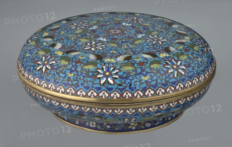 Box with lid, gift, Chinese, Qing dynasty Qianlong period, cloisonné enamel. Large cylindrical-spheroidal box supported on a separate annular foot, with a slightly flattened lid. Covered by a plant tendril encircling the entire surface container and lid, dominated by chrysanthemum flowers and leaves in various stylistic forms; rich multicolored plant composition on a turquoise background. Upper edge of the storage section and lower edge of the cover decorated with a border of ruyi-head motifs in white and red; a blue three-petal pattern completes the decoration. Foot decorated with an ornamental band of colorful flowers and leafy twigs. Interior of the box and inner surface of the foot filled with uniform turquoise enamel. Unglazed edges of the storage section, cover, and foot gilded. 18th century, China. Height 14.5 cm, diameter 34.5 cm., A rounded decorative box with a domed lid covered in an intricate floral and scroll pattern in shades of blue, turquoise, green, white, yellow, brow...