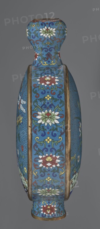 Vase - moon bottle pilgrim bottle with a flattened belly, supported by a separate oval high foot, with a short neck resembling a garlic bulb suantoukou type. Decoration on one side: a multicolored composition depicting a chrysanthemum flower growing on a coastal rock, above which a dragonfly and a grasshopper hover. The other side shows lilies irises on a rock above the water, with a butterfly and a grasshopper above them. Small colorful flowers growing on the shore appear in both representations. Both compositions use the flowers and birds motif known from Chinese scroll painting. A Chinese meander forms the background for both compositions. The foot, side edges, and neck are filled with a lotus tendril pattern. Colors dominated by red, white, green, yellow, and navy blue; elements without enamel decoration are gilded. Visible abrasions of the gilding and technological firing defects. Cloisonné enamel. Qing dynasty Qing style, 2nd half 18th century, China. Height 29.8 cm, width 21.1 c...