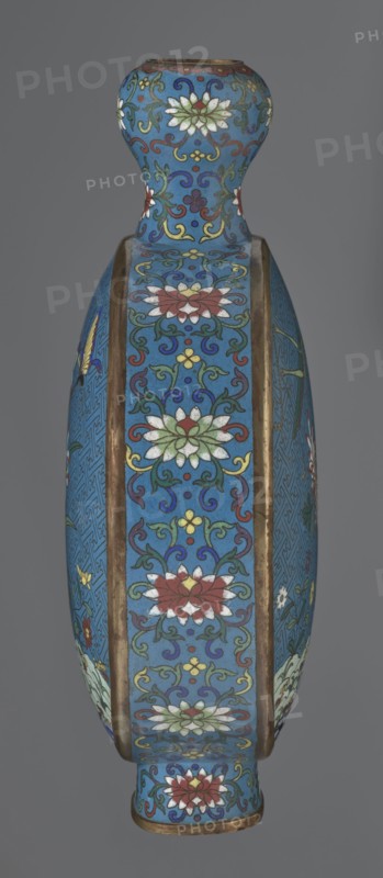 Vase - moon bottle pilgrim bottle with a flattened belly, supported by a separate oval high foot, with a short neck resembling a garlic bulb suantoukou type. Decoration on one side: multicolored composition depicting a chrysanthemum growing on a coastal rock, above which a dragonfly and a grasshopper hover. Other side: composition showing lilies irises on a rock above the water, with a butterfly and a grasshopper; small colorful flowers growing on the shore visible in both representations. Motif derives from Chinese flowers and birds scroll painting. A Chinese meander forms the background for both compositions. Foot, side edges, and neck filled with a lotus tendril; palette dominated by red, white, green, yellow, and navy blue. Elements without enamel are gilded; visible abrasions of the gilding and firing-related technological defects. Cloisonné enamel. Qing dynasty 1644–1911, 2nd half 18th century. Chinese. Vessel. Height 29.8 cm; width 21.1 cm., A tall vase with a rounded shoulder a...