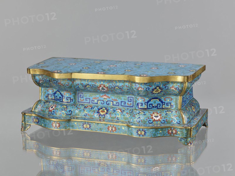 Wall pedestal; creator, cloisonné enamel; decoration: flowers on a turquoise background with a grid of cracks cracked ice, floral tendrils on the profiles, and a central band with geometric motifs; plant and floral and geometric ornament; Qing dynasty 1644–1911, 18th century; China; height 13.5 cm, width 36.6 cm, depth 18.5 cm., A small ornate rectangular box with a wavy, scalloped profile sits on a reflective surface, showing a mirrored underside. The box is covered in a bright turquoise-blue enamel background with intricate floral and foliate patterns in colors including blue, red, white, green, and gold, with gold bands edging the top and base. The sides feature repeating decorative panels with stylized geometric motifs and scrolling vinework, and the top surface displays scattered floral rosettes set against a network of fine crackle lines. The form includes curved, bulging sections and small bracket feet, and the overall appearance is glossy and richly decorated.