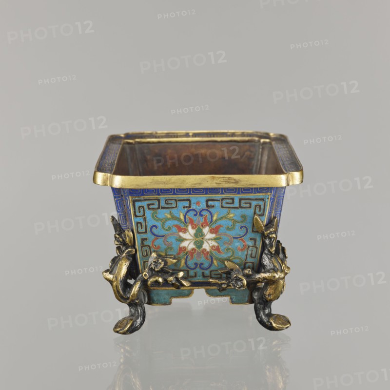 Flower pot incense burner on four legs, 18th century, China, Qing 1644–1911. Cloisonné enamel vessel, jardinière/incense burner; decoration: lotus tendril on a turquoise background, legs shaped as flowered trunks plum branches made of gilded bronze; motifs include lotuses, lotus tendril, plum trees, cracked ice, and meander ornament. Dimensions: height 9.5 cm, width 17.2 cm, depth 12.0 cm., A small square metal container with a flared gold rim sits against a plain light gray background; its exterior surfaces are decorated with a turquoise blue base and intricate patterns in darker blue, green, red, white, and gold forming a stylized floral motif on the visible side. The container rests on ornate feet shaped like twisted branches and animal-like forms in dark metal with gold highlights. The interior appears brown and worn, while the exterior corners show geometric border patterns in blue and gold. The overall colors visible include turquoise blue, dark blue, green, red, white, gold, dar...