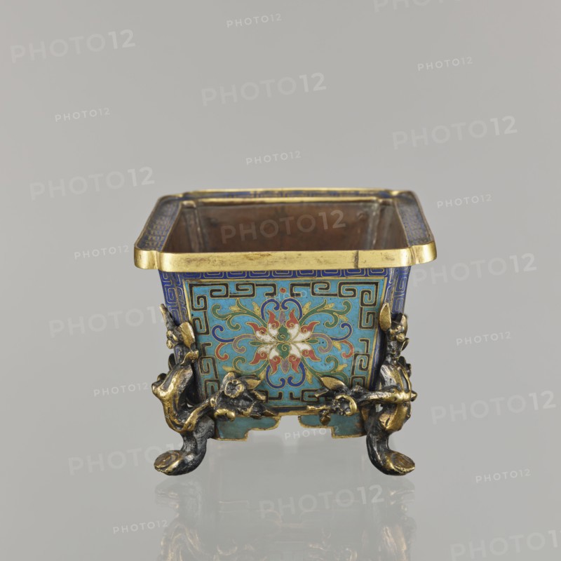 Flower pot incense burner on four legs, cloisonné enamel; decoration: lotus tendril on a turquoise background; legs in the shape of flowered trunks plum branches made of gilded bronze; motifs include lotuses, plum trees, cracked ice, meander; jardinère form; 18th century, Qing 1644–1911, China; vessel; height 9.5 cm, width 17.2 cm, depth 12.0 cm., A small square metal container with an open top sits on a reflective surface, showing a decorative exterior in blue, turquoise, gold, red, white, and green. The upper rim is gold, and each side panel features a central floral motif with swirling petal shapes in red, white, and green set against a turquoise background framed by blue geometric borders. Ornate gilt and dark metal mounts form a group of sculpted elements at the corners and act as feet with curved, claw-like ends. The interior of the container appears plain and metallic with a brownish tone. The background is a uniform light gray.