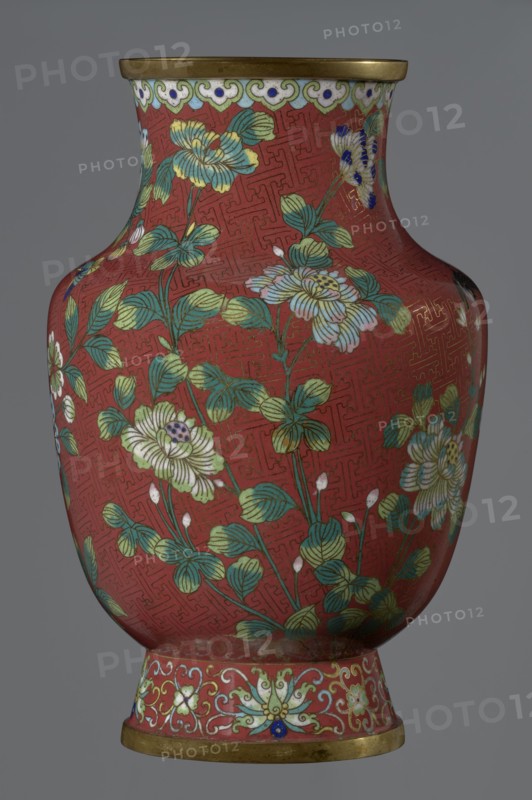 Vase, Chinese art; Chinese culture. Qing dynasty 1644–1911, Xianfeng period 1851–1861. Cloisonné enamel with floral pattern and meander ornament. The vase is set on a tall, conical foot flaring downwards and terminating in a metal ring, decorated with a lotus tendril. The neck is short and wide, topped with a metal ring, below which is a border composed of ruyi heads. The belly and neck are decorated with a continuous composition of flowers and plum branches against a red Chinese meander background. 1st half of the 19th century. China. Vessel. Height 25.1 cm; diameter 16.5 cm; weight 1.5 kg., A decorative vase photographed against a plain gray background, showing its rounded body, narrowed neck, and flared rim and foot with metallic bands. The main surface of the vase is a deep red color with an intricate repeating geometric pattern in a slightly darker shade. Over this red ground there is a floral design featuring a few large blossoms and many leaves in shades of green, pale yellow, w...