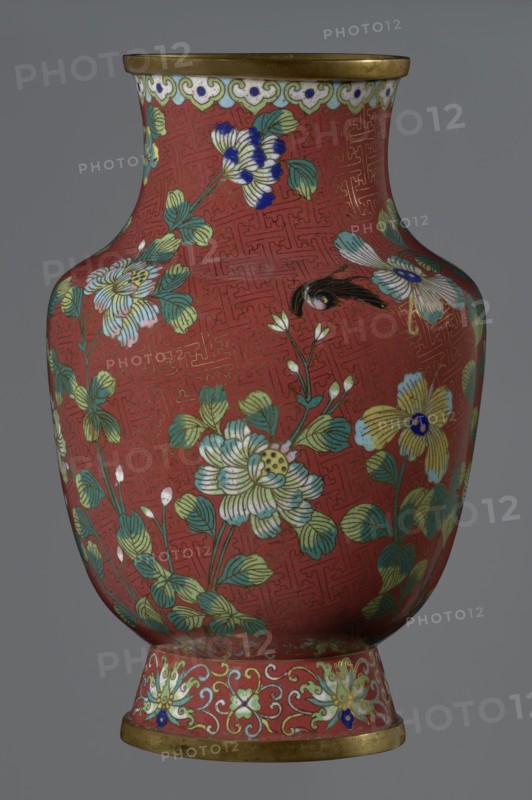 Vase, cloisonné enamel; Chinese, Qing dynasty, Xianfeng period 1851–1861; 1st half of the 19th century; height 25.1 cm, diameter 16.5 cm, weight 1.5 kg. The vase is set on a tall, conical foot flaring downwards and terminating in a metal ring, decorated with a lotus tendril. The short, wide neck is topped with a metal ring beneath which is a border composed of ruyi heads. The belly and neck are decorated in a continuous composition of flowers and plum branches against a red Chinese meander background., A rounded vase with a narrow neck and flared rim sits against a neutral background, its main surface a deep red decorated with an intricate repeating geometric pattern in a slightly darker red. A band near the rim shows a scalloped cloud motif in pale green, white, and blue with gold accents, and the rim itself is a metallic gold color. The body of the vase is covered with floral designs featuring large stylized blossoms and leaves in shades of pale green, light blue, cream, and yellow, ...