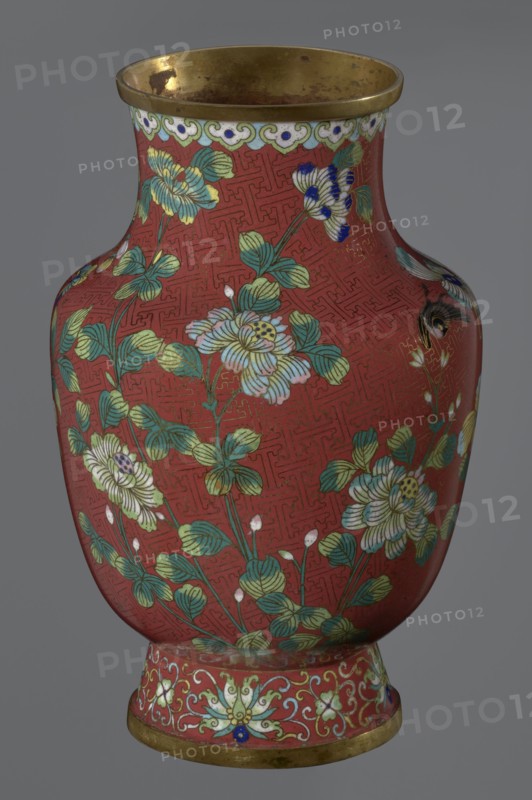 Vase, noted; Chinese art; Qing dynasty 1644–1911, Xianfeng period 1851–1861; cloisonné enamel; floral motifs and meander ornament. The vase is set on a tall, conical foot flaring downwards and terminating in a metal ring, decorated with a lotus tendril. The neck is short and wide, also topped with a metal ring, below which is a border composed of ruyi heads. The belly and neck are decorated with a continuous composition of flowers and plum branches against a red Chinese meander background. 1st half of the 19th century; China; vessel. Height 25.1 cm, diameter 16.5 cm, weight 1.5 kg., A rounded vase with a flared rim and a slightly footed base sits against a neutral gray background; the vase surface is predominantly red with an allover geometric pattern and is decorated with a group of stylized flowers and leaves in shades of green, teal, pale yellow, white, and light blue, with some flower accents in dark blue and black; the rim and base edges are metallic gold and show some darker spot...