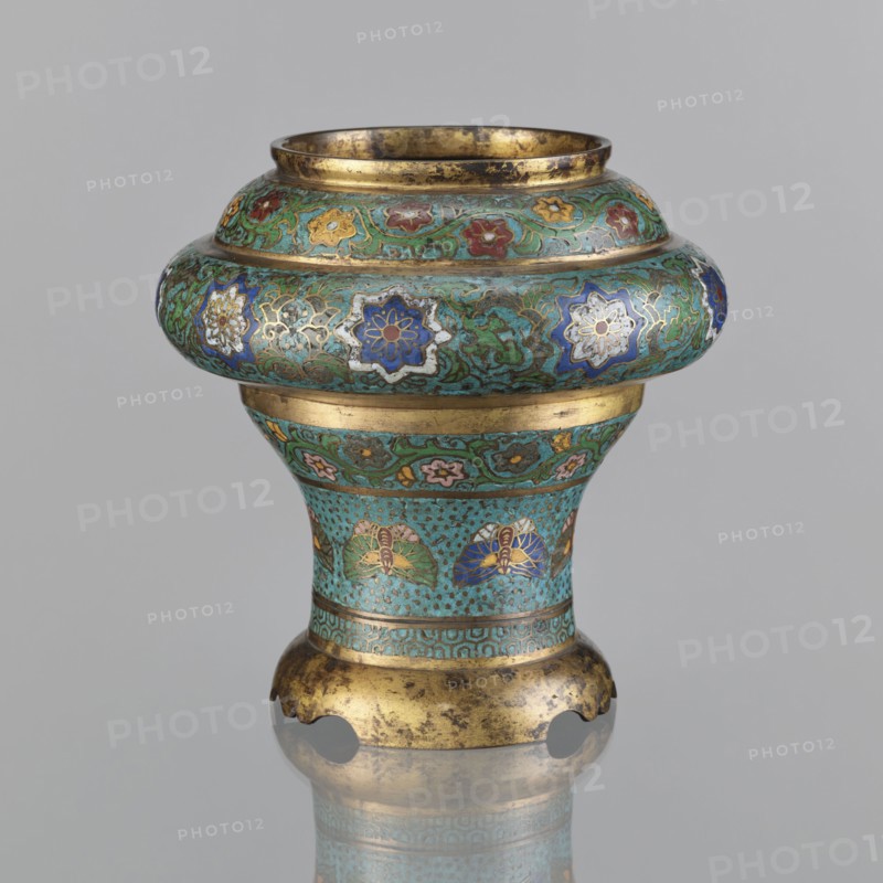 Vessel, Japanese, Meiji period 1868–1912, 19th century. Materials/technique: champlevé enamel. Decoration: plant motifs, butterflies, flowers, fans; sasarindo coat of arms. Dimensions: height 28 cm; diameter 28.6 cm., An ornate vessel with a rounded upper section narrowing into a flared base sits against a plain light gray background; its surface shows a turquoise green patina with intricate floral and vine motifs in shades of blue, white, red, yellow, pink, and green, accented with metallic gold bands and gilt areas that show darker wear; the upper rim and base edges are gold-toned with irregular dark spots, and the piece displays repeating stylized flower medallions and leaf-like patterns around the midsection, with a reflective surface beneath suggesting a faint reflection.