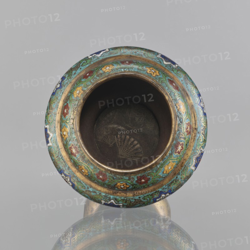 Vessel, Meiji period 1868–1912, Japan. 19th century. Champlevé enamel decoration with plant motifs, butterflies, flowers, and fans; sasarindo coat of arms. Height 28 cm; diameter 28.6 cm., A round ornate vessel rim viewed from above showing an open dark interior with faint engraved fan shapes at the bottom, surrounded by a wide decorative band featuring a repeating floral and scroll pattern in turquoise green, blue, red, yellow, white, and gold, with a visible reflective surface beneath that mirrors parts of the vessel; the background is a uniform light gray.
