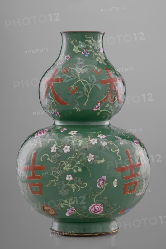 Vase, calabash form, 18th century, Qing dynasty, China. Porcelain/ceramic vessel, height 34.3 cm, diameter 24.0 cm. Decoration on a green background with flowers, butterflies, fruit peaches, lingzhi, peonies and floral ornament; pictographic signs among the plant motifs. Chinese art; Qing style., A double-gourd porcelain vase with a glossy green ground decorated with floral vines, blossoms, and berries in colors including pink, red, purple, blue, and gold, and featuring several bold red graphic shapes on both bulbous sections; the vase has a narrow neck, a flared rim with a thin brown edge, and a green foot with a thin brown band at the base, all set against a neutral gray background.