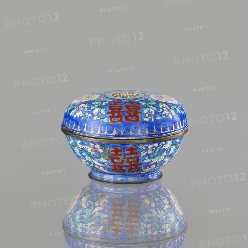 Incense burner - bowl with a lid, Chinese, Qing dynasty 1644–1911, 18th/19th century. Painted enamel decoration on a navy blue background with flower tendrils peony, bats, flowers, coins, and the character shuangxi double happiness, usually used in China in the context of marriage. Ceremonial vessel — censer/incense burner. Height 6.4 cm; diameter 9.7 cm; weight 0.17 kg., A small round lidded box sits centered against a plain light gray background, showing a glossy surface and a clear reflection beneath it. The box is predominantly bright blue with decorative floral patterns in shades of green, pink, white, and pale purple, and has ornate gold accents along the rim and base. There are bold rectangular motifs in deep red on both the lid and the body, and the overall finish appears enamel-like and polished.