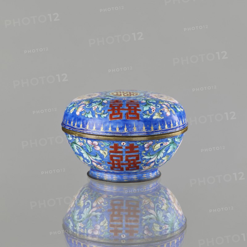 Incense burner - bowl with a lid, Chinese art, Chinese, Qing 1644–1911 style, bats iconography, ceremonial vessel, plant ornament, censers, flowers, coins, painted enamel, happiness symbol, export product, decoration on a navy blue background, apart from the flower tendril — peony, the character shuangxi double happiness — usually used in China in the context of marriage, 18th/19th century, China, vessel, incense burner, height 6.4 cm, diameter 9.7 cm, weight 0.17 kg, A small rounded box with a domed lid sits centered on a reflective surface, showing its mirror image below. The box is predominantly bright blue with decorative floral patterns in shades of green, pale pink, and white, and has accents of gold along the rim and base. There are bold red geometric motifs on both the lid and the body. The overall shape is circular with a slightly flared foot, and the background behind the object is a plain neutral gray.