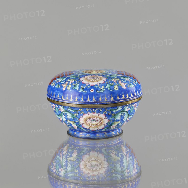 Incense burner — bowl with a lid; maker, ; Chinese; Qing dynasty 1644–1911; 18th–19th century; painted enamel on metal; decoration on navy blue background with flower tendrils peony, bats, flowers, coins; character shuangxi double happiness — usually used in China in the context of marriage; ceremonial vessel / censer; export product; height 6.4 cm, diameter 9.7 cm, weight 0.17 kg., A small rounded decorative box with a domed lid sits centered on a reflective surface, the box predominantly featuring a bright cobalt blue background with floral and foliate patterns in pink, green, turquoise, white, and gold; the lid and body each display a large stylized blossom surrounded by curling green leaves and smaller pink flowers, with gold accents and a thin gold band where lid meets base, and the box’s reflection mirrors the same colors and motifs on the grayish surface beneath.