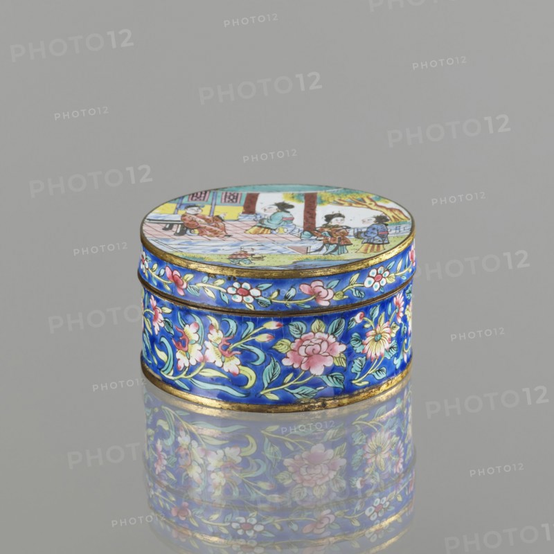Box with a lid;, painted enamel; Qing dynasty 1644–1911; 19th century; Vietnamese Nguyen dynasty, 1802–1945 and Chinese cultural influence; figurative scenes depicting peonies, grapes, and peaches iconography; container; height 3.3 cm, diameter 6.3 cm., A small round box with a domed lid sits on a reflective surface, showing a bright palette of blue, pink, green, yellow, white, gold, and brown. The box exterior is predominantly cobalt blue with painted floral motifs in pink, white, and yellow with green leaves and gold accents that form a decorative band around the sides. The lid features a painted scene with small figures and architectural elements rendered in muted tones of yellow, brown, blue, green, and pink, bordered by a thin gold rim. The reflection of the box on the surface repeats the blue and floral patterns and soft colors of the lid.