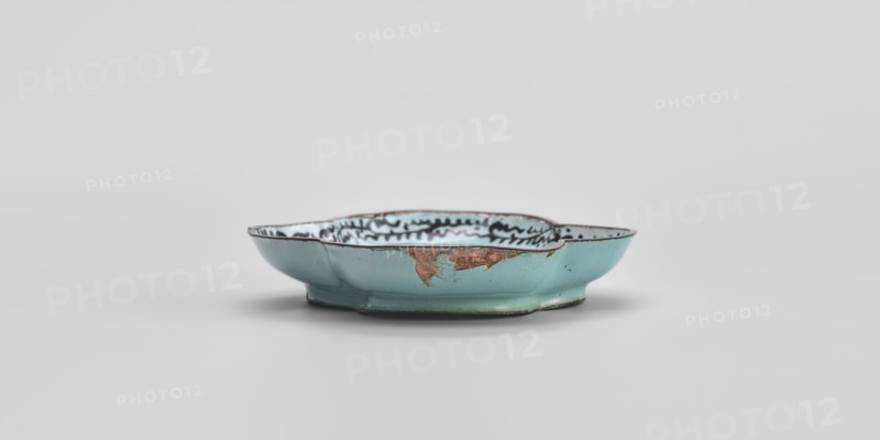 Plate, painted enamel on vessel, Chinese, Qing 1644–1911, 18th century, genre scenes, height 1.3 cm, width 7.5 cm, depth 9.1 cm., A shallow, irregularly shaped ceramic dish sits centered against a pale gray background; the dish is predominantly turquoise on its exterior with areas of exposed brownish material and fine hairline cracks, and its interior shows a lighter bluish-white glaze with a dark wavy decorative pattern near the rim; the rim itself appears darker with subtle wear, and the dish casts a soft shadow beneath it.