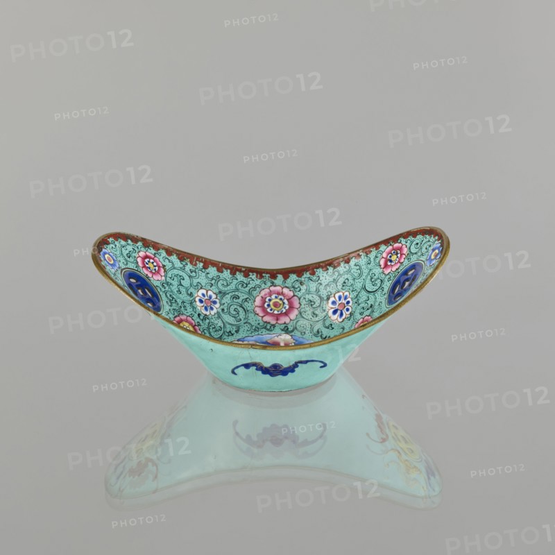 Bowl; painted enamel on porcelain; Qing dynasty 1644–1911, 18th–19th century, China; decorated with lotuses, coins, flower tendrils and bats symbolizing happiness; vessel height 6.3 cm, width 8.5 cm, depth 15.3 cm., A shallow, crescent-shaped decorative bowl sits on a reflective surface, showing a mirrored image beneath. The bowl's exterior is a pale turquoise with a dark blue stylized motif on the front, while the interior displays an intricate patterned background in a slightly darker turquoise filled with swirling designs and floral medallions in pink, white, and blue, each with contrasting centers. The interior rim has a narrow band of rusty red edged with a thin gold border, and there are additional circular blue medallions set into the patterned interior. The overall color palette includes shades of turquoise, pink, blue, white, rusty red, gold, and small accents of darker tones.