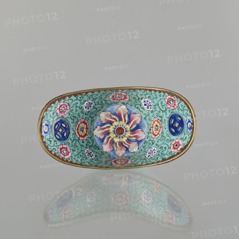 Bowl, painted enamel, Qing 1644–1911, China, 18th/19th century; decorated with boats, lotuses, coins, flower tendril, and bats iconography; symbolizing happiness; vessel; height 6.3 cm, width 8.5 cm, depth 15.3 cm., An oval decorative dish with a gold rim sits against a plain light gray background and casts a soft reflection below; the interior surface is covered in an intricate turquoise-green pattern with swirling lines and features a central circular floral motif in blue, pink, yellow, and red surrounded by several smaller flower designs in pink, blue, yellow, and white, as well as a few circular blue medallions.