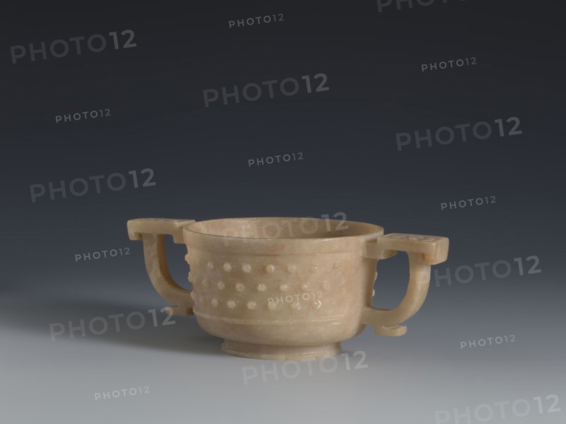 Gui-type bowl, Chinese, Qing dynasty 1644–1911, 2nd half 18th century. Porcelain vessel modeled after a ritual gui. Supported by a low ring foot; walls decorated with three horizontal rows of small convex knobs evoking archaic ruding nailheads, framed by a thin rim at the bottom and a band framing the spout at the top. Two stylized handles on either side reference the ancient prototype. Upper part decorated with an image of cicada larvae arranged in a rosette. Height 4.5 cm; width 13 cm; diameter 7.2 cm., A shallow, round cup-like vessel with a warm beige color sits centered on a smooth surface, featuring a band of raised rounded bumps around its exterior; it has a short pedestal base and two curved handles extending outward on opposite sides that are the same beige tone, and the background transitions from a dark gray at the top to a lighter gray near the surface.