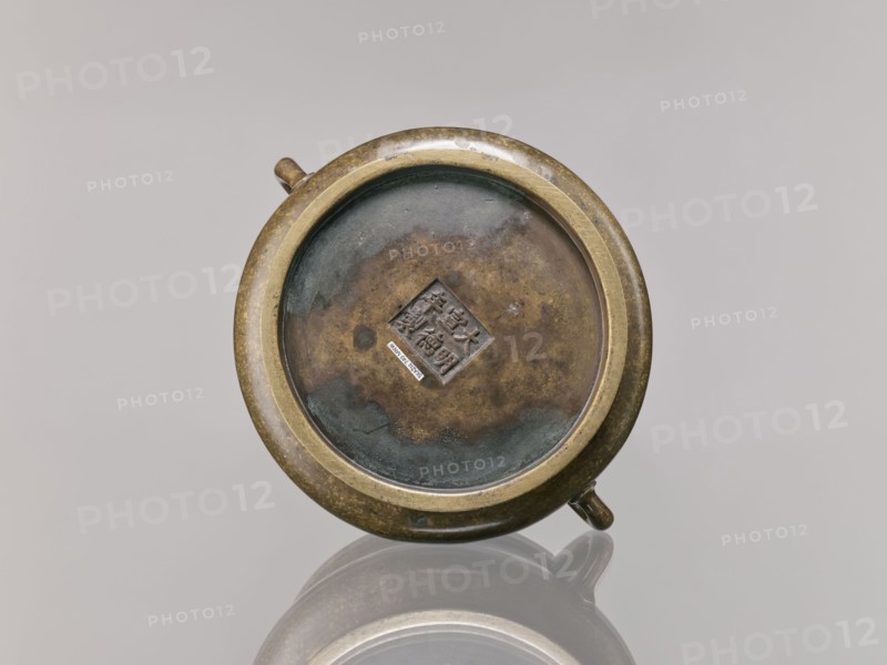 Censer, Chinese, Qing dynasty 1644–1911, 18th century; ceremonial/ritual vessel; backdated signatures; bronze censer; height 8.5 cm, diameter 16.8 cm., A round metal object with a circular rim and a recessed center sits against a plain light gray background, showing a patina of brown, green, and dark bluish-gray tones on its surface; the rim appears golden-bronze with areas of darker mottling, and there are a few small protruding knobs on the outer edge; the object casts a soft reflection on the glossy surface below, and the overall scene includes shades of brass, brown, green, bluish-gray, and light gray.