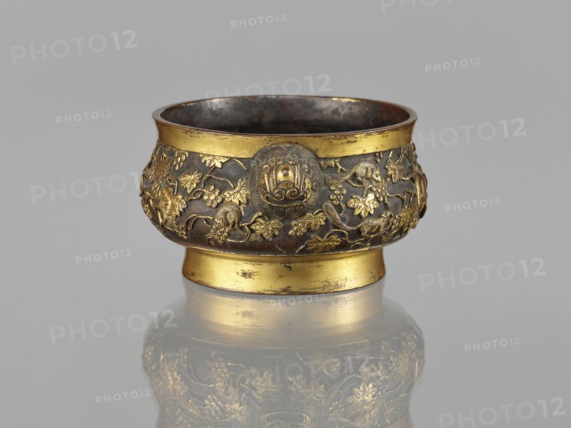 Gui-type incense burner, Li, Daguan fl. late 16th–early 17th century? signature, Chinese art, Chinese culture, Qing 1644–1911, zoomorphic motifs, gui vessels, plant ornament, ceremonial vessel, squirrels, vines, lions iconography, grapes, handles, ritual vessel, 18th/19th century, China, vessel, incense burner; height 6.5 cm, diameter 11.7 cm, weight 0.56 kg, A small ornate bowl with a rounded body and a short pedestal base sits against a light gray background, showing metallic surfaces in gold and dark brown; the upper and lower rims are gold while the midsection is dark brown with raised gold decorative motifs depicting a dense pattern of leaves, vines, and animal figures, and a round medallion-like ornament on the front; the interior appears dark and slightly reflective, and the object casts a soft reflection on the surface beneath it.