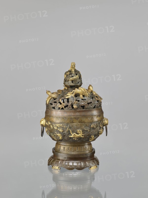 Censer with a cover on a base, maker, Qing style, Chinese art, Chinese culture; ceremonial/ritual vessel, censer with a high foot. Openwork cover and handles. Decoration: dragons and hôô phoenix birds on the cover; various animals tigers, birds, fish, phoenixes on the belly; dragon with a pearl, sea waves, clouds, geometric patterns, meander ornament. Cover with removable knob. 18th century, China. Dimensions: height 22.0 cm, width 21.0 cm, depth 17.7 cm., An ornate metal vessel sits on a rounded pedestal base, showing a mix of brown, bronze, and gold tones. The upper portion has a perforated domed lid with decorative cutouts and sculpted elements, including a finial at the top. A group of sculpted motifs, some raised and gilded, decorate the lid and shoulder, with curving, organic shapes and small animal-like forms. The body of the vessel displays a band of patterned engraving below the perforated section and a lower area with textured relief scenes and gilded decorative figures. Two ...