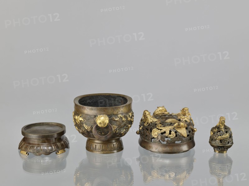 Censer with a cover on a base;, Qing period 18th century, China. Bronze vessel censer with high foot, openwork cover and handles; cover has removable knob. Decoration: dragons and ho-o phoenix birds on the cover; various animals on the belly including tigers, birds, fish; motifs of dragon with a pearl, clouds, sea waves, geometric patterns meander. Ritual/ceremonial vessel. Height 22.0 cm, width 21.0 cm, depth 17.7 cm., A group of small metal objects arranged in a row on a reflective surface against a neutral gray background: a low round container with a ridged base and a flat lid in dark bronze tones with some golden accents; a larger bowl-shaped vessel with a wide rim, ornate relief decoration and a dark interior, showing bronze and gilded areas; a domed, openwork cover richly decorated with sculpted creatures and floral forms in mixed bronze and bright gold; and a smaller ornate finial-like piece with openwork decoration and gilded detailing. The visible colors include bronze, dark ...